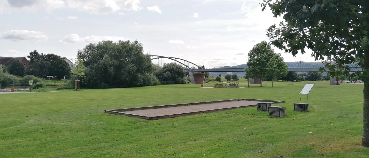 Boulebahn Beverungen Boulebahn auf großer Rasenfläche mit Sitzwürfeln und Informationstafel vor der Kulisse der Weserbrücke.