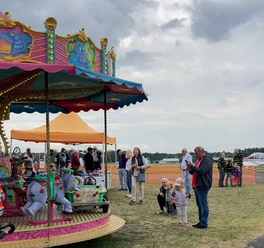 Buntes Kinderkarussell mit Elefanten, umgeben von Familien auf einer Wiese unter bewölktem Himmel.