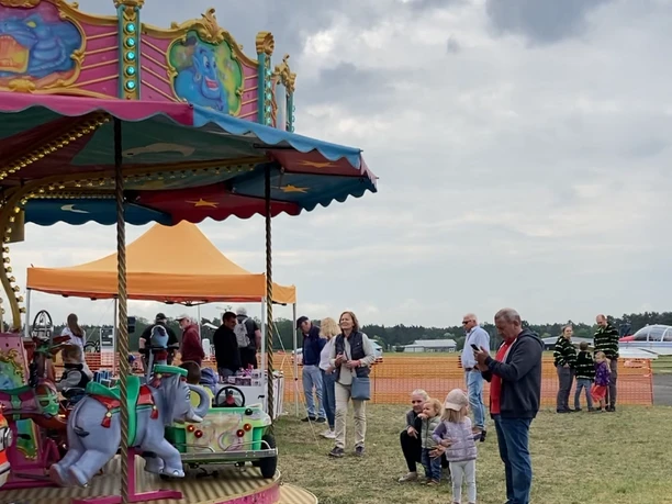 Buntes Kinderkarussell mit Elefanten, umgeben von Familien auf einer Wiese unter bewölktem Himmel.
