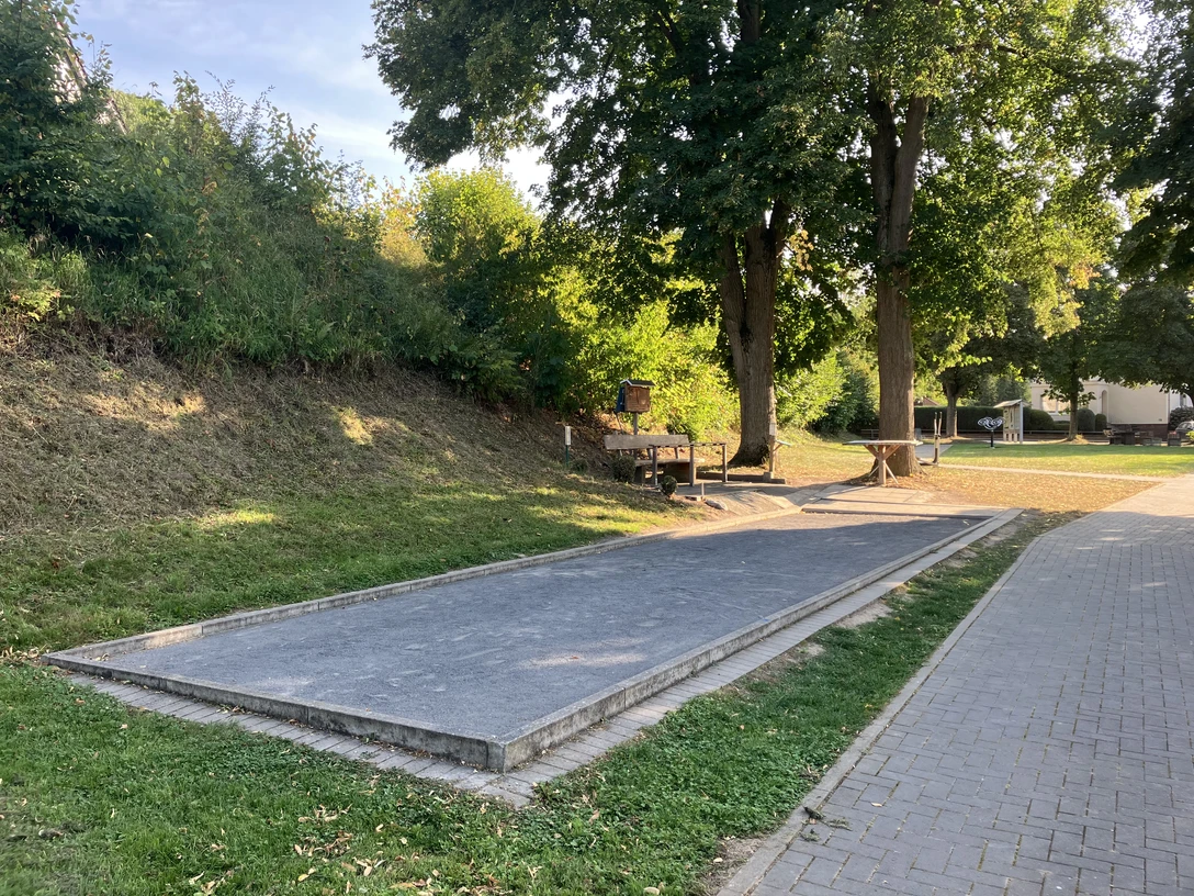 Eine Boulebahn mit Sitzgelegenheiten unter zwei großen Bäumen.