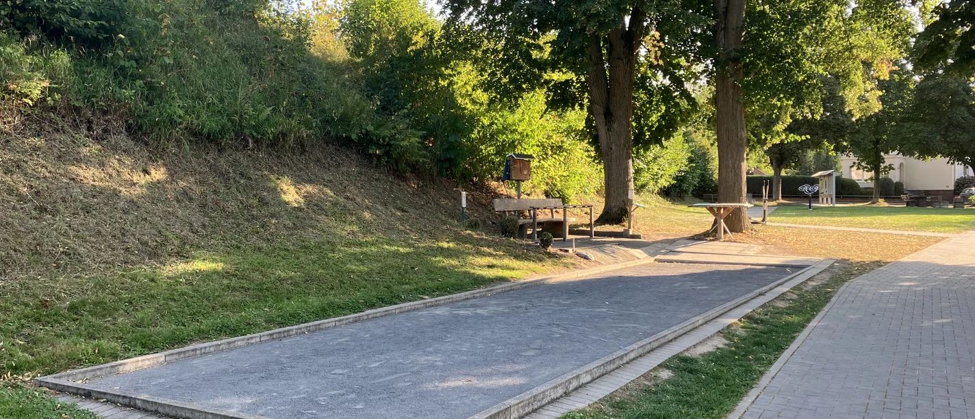 Eine Boulebahn mit Sitzgelegenheiten unter zwei großen Bäumen.