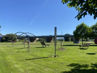 Bewegungsparcours Beverungen Auf einer Wiesen stehen sechs verschiedene Bewegungsgeräte und Liegebänke, im Hintergrund befindet sich die Beverunger Weserbrücke.