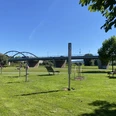 Bewegungsparcours Beverungen Auf einer Wiesen stehen sechs verschiedene Bewegungsgeräte und Liegebänke, im Hintergrund befindet sich die Beverunger Weserbrücke.