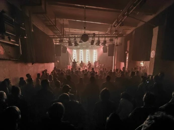 Gebäude 9 Das Bild zeigt eine gut besuchte Konzertveranstaltung in einem düsteren Raum mit stimmungsvoller Beleuchtung.The picture shows a well-attended concert event in a gloomy room with atmospheric lighting.