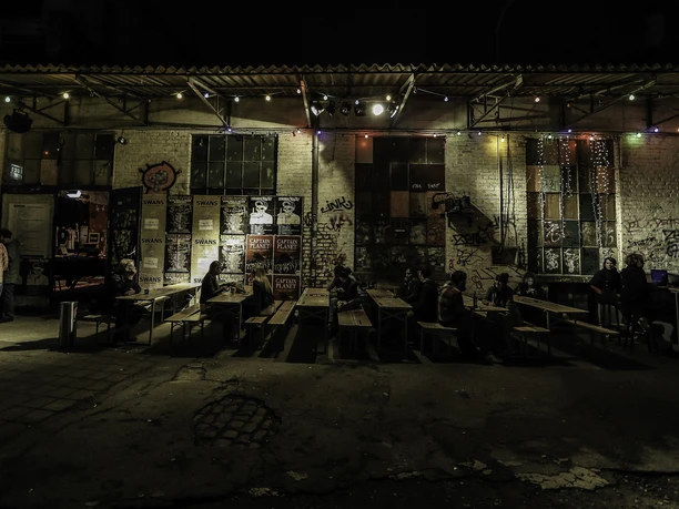 Gebäude 9 Colorful fairy lights illuminate an exterior façade while people sit at rustic wooden tables.