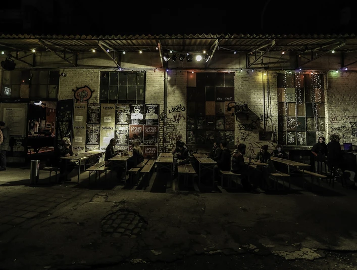 Gebäude 9 Bunte Lichterketten erhellen eine Außenfassade, während Menschen an rustikalen Holztischen sitzen.Colorful fairy lights illuminate an exterior façade while people sit at rustic wooden tables.
