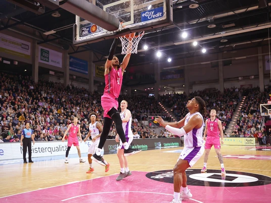 Telekom Baskets Bonn Spieler in pinken Trikots dominieren ein Basketballspiel in einer großen, voll besetzten Arena.Players in pink jerseys dominate a basketball game in a large, packed arena.