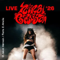 Eine Künstlerin performt live auf einer Bühne, umgeben von dramatischem Rauch und rotem Schriftzug.