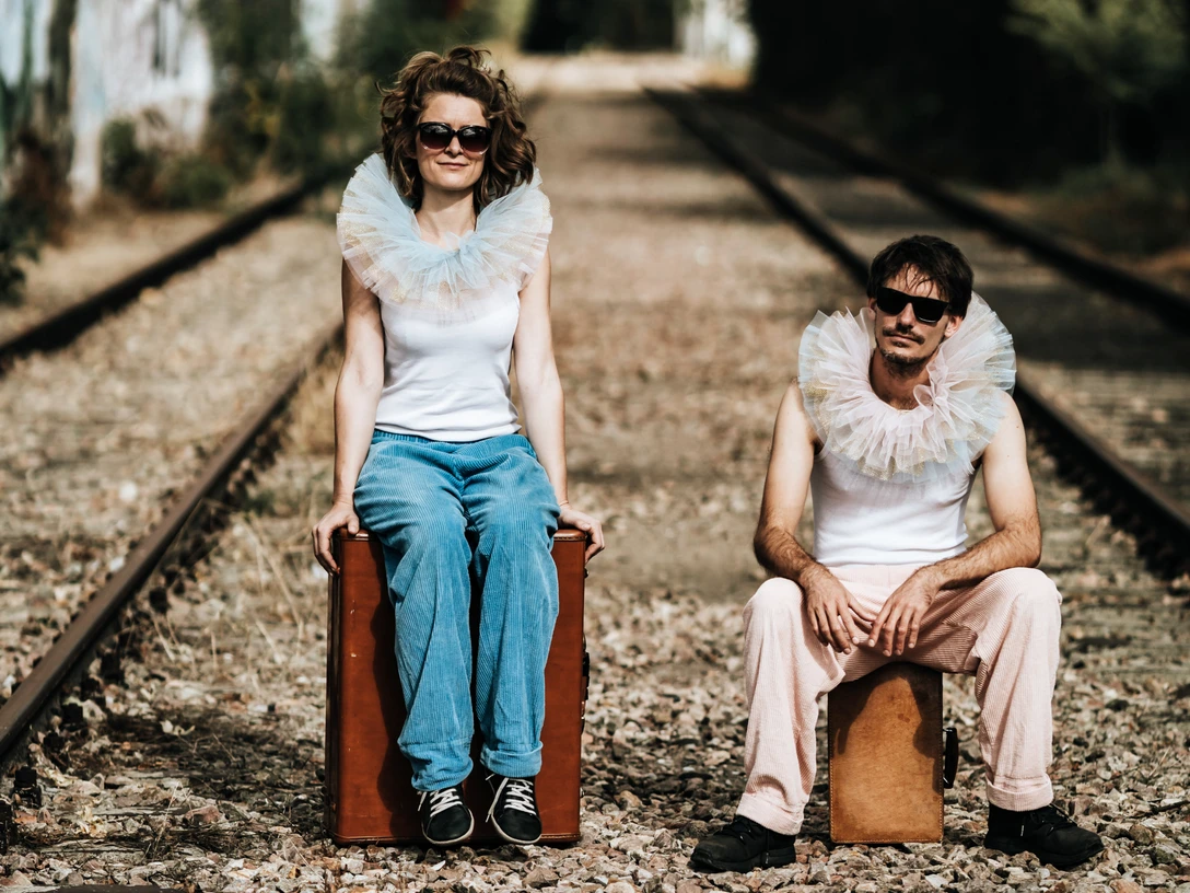 MF-Presse-07-Fotograf-Max-Saufler.jpg Zwei Personen mit Tüllkrägen und Sonnenbrillen sitzen auf Holzkisten, umgeben von Bahngleisen.
