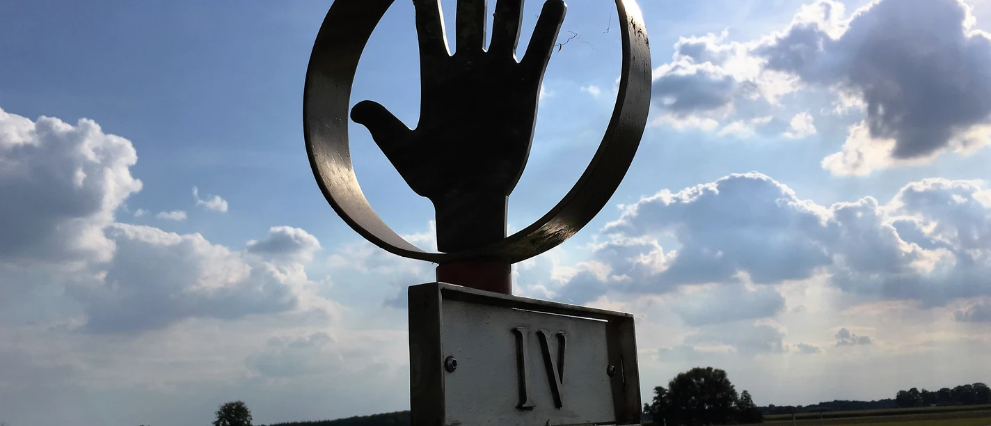 Römerlager Delbrück-Anreppen Skulptur einer Hand in einem Kreis vor einem weiten Feld, Wolken und blauem Himmel im Hintergrund.