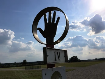 Römerlager Delbrück-Anreppen Skulptur einer Hand in einem Kreis vor einem weiten Feld, Wolken und blauem Himmel im Hintergrund.