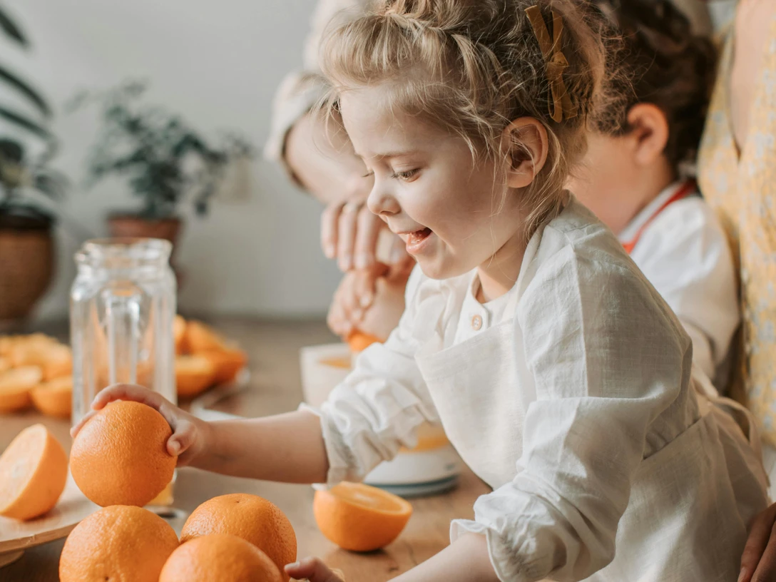 pexels-vlada-karpovich-4617285.jpg Ein begeistertes Kind greift nach Orangen, während Eltern im Hintergrund lächelnd zusehen.