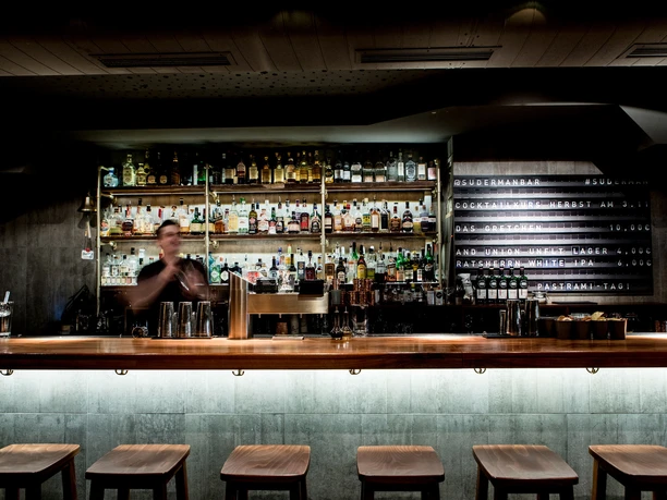 Suderman A bartender mixes drinks in front of a wall of shelves with various spirits in a bar.