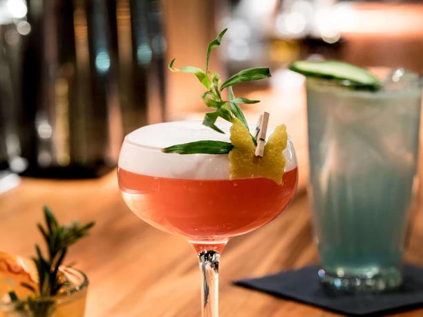 Suderman A cocktail glass with red liquid and a head of foam stands on a wooden counter.