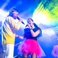 260207-batch_eighties-kamen--7074.jpg Zwei Musiker im bunten Licht einer Bühne, mit Mikrofon und Saxophon, singend und spielend.