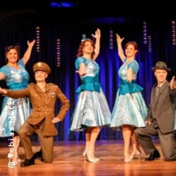 Vier Personen, zwei in blauen Kleidern und zwei in Uniform, stehen lächelnd auf einer Theaterbühne.
