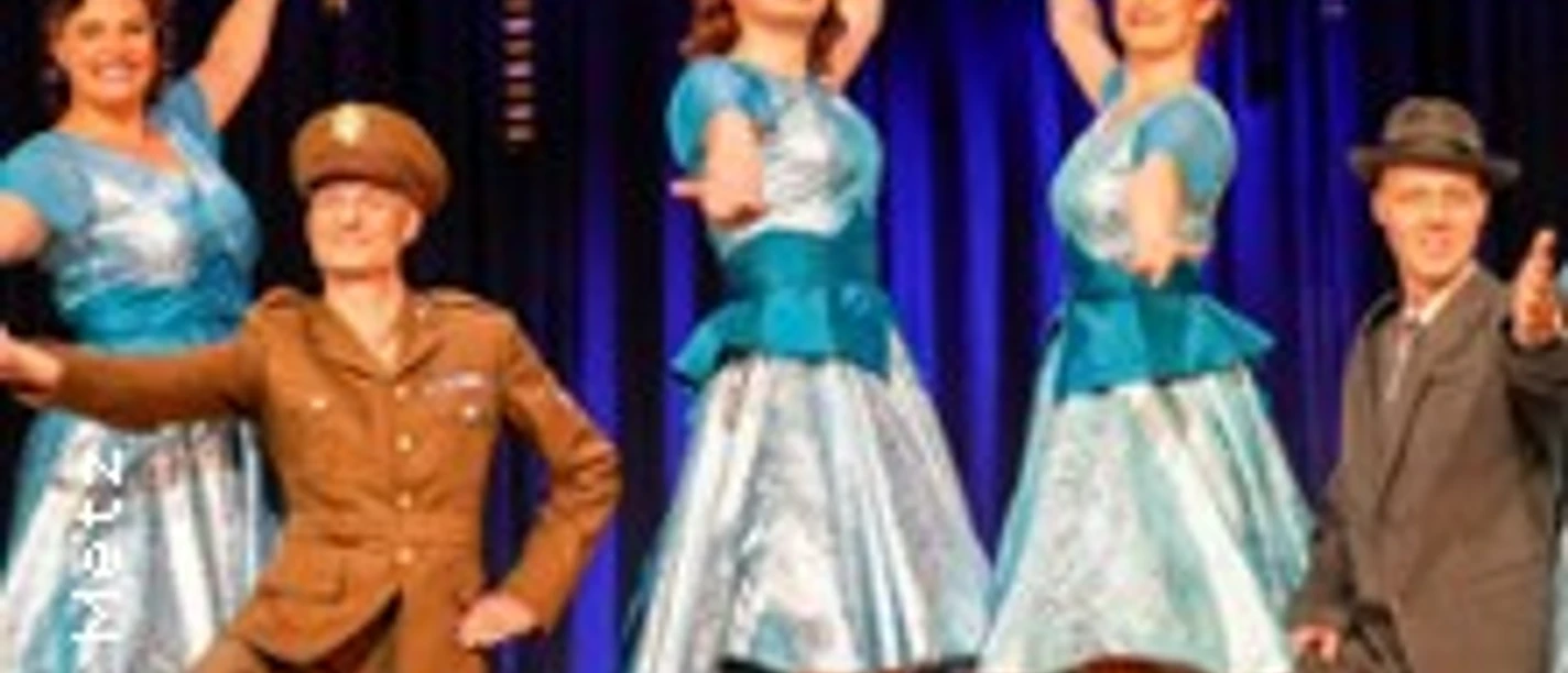 Vier Personen, zwei in blauen Kleidern und zwei in Uniform, stehen lächelnd auf einer Theaterbühne.