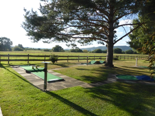 Minigolfanlage Dreiländereck in Beverungen Mehrere Minigolfbahnen auf einem umzäunten Rasengrundstück unter einer großen Konifere.