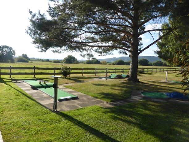 Minigolfanlage Dreiländereck in Beverungen Mehrere Minigolfbahnen auf einem umzäunten Rasengrundstück unter einer großen Konifere.