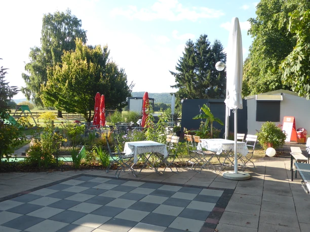 Kiosk mit Sitzmöglichkeiten, Sonnenschirmen und einem Schachspielfeld, umrahmt von einzelnen Bäumen.