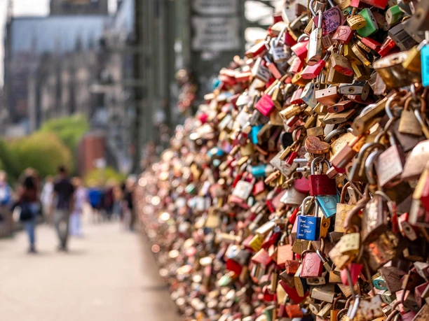 Love locks in Cologne A multitude of colorful love locks adorn the railing of the Hohenzollern Bridge in Cologne. The silhouette of the majestic Cologne Cathedral blurs in the background, complemented by the people passing by on the bridge as they cross the Rhine on foot.