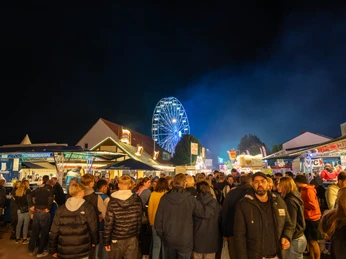 Gesmolder Kirmes.jpg Menschenmenge auf einem belebten, abendlichen Jahrmarkt mit beleuchtetem Riesenrad im Hintergrund.