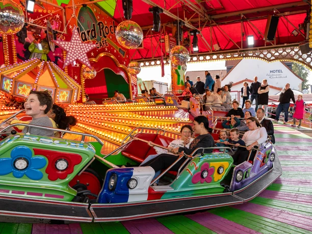 Gesmolder Kirmes.jpg Menschen fahren auf einem bunten Karussell auf einem Jahrmarkt unter einem roten Zeltdach.