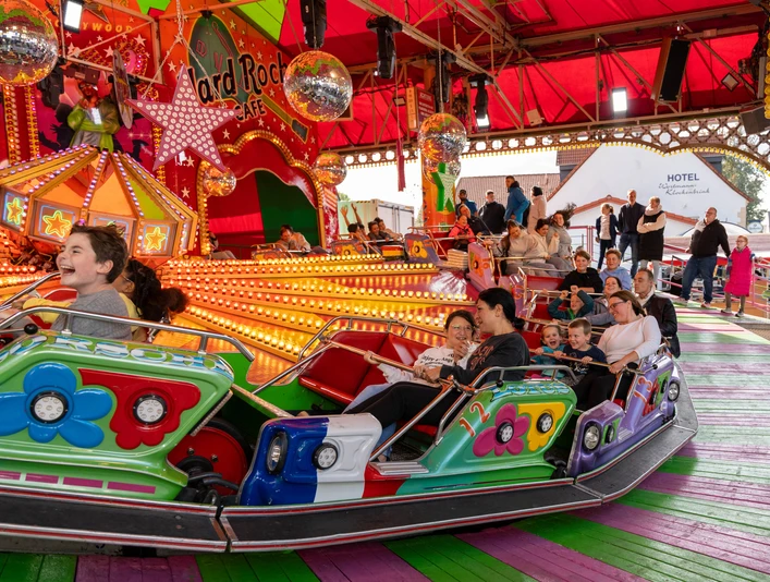 Gesmolder Kirmes.jpg Menschen fahren auf einem bunten Karussell auf einem Jahrmarkt unter einem roten Zeltdach.