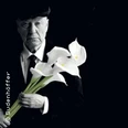 Ein älterer Mann in Anzug hält weiße Calla-Lilien, vor dunklem Hintergrund in fokussierter Pose.