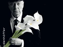 Ein älterer Mann in Anzug hält weiße Calla-Lilien, vor dunklem Hintergrund in fokussierter Pose.