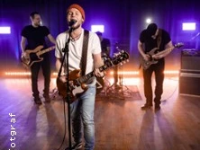 Vierköpfige Band auf einer Bühne spielt mit Gitarren und Schlagzeug vor leuchtenden Neonlichtern.