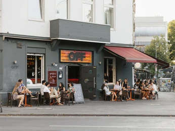 OFF Bar Vor der grauen Fassade der OFF-Bar in Köln sitzen Menschen entspannt unter einem roten Sonnensegel.People sit relaxed under a red awning in front of the gray façade of the OFF-Bar in Cologne.