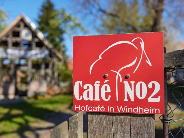 Café No2 mit CaféGarten | Historische Hofstelle Windheim No2 im Mühlenkreis Minden-Lübbecke Rotes Schild für das Hofcafé No2 in Petershagen, mit Hofstelle im Hintergrund bei Sonnenschein.