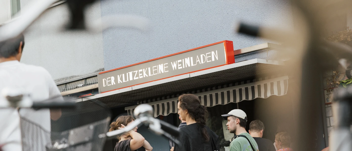 Der klitzekleine Weinladen Vor einem kleinen Geschäft in der Stadt versammeln sich Menschen. Fahrräder sind im Vordergrund.