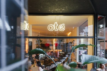 Oté Das Bild zeigt die helle und einladende Eingangsfassade eines Cafés oder Restaurants mit großen Glasfenstern und stilvoller Inneneinrichtung.