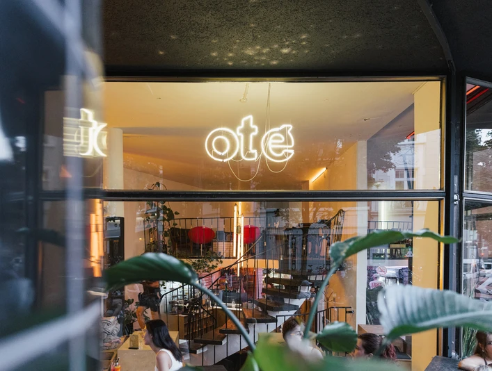 Oté Das Bild zeigt die helle und einladende Eingangsfassade eines Cafés oder Restaurants mit großen Glasfenstern und stilvoller Inneneinrichtung.