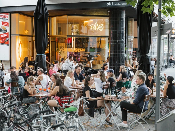 Oté A crowd enjoys drinks outside a café with large windows in Cologne.