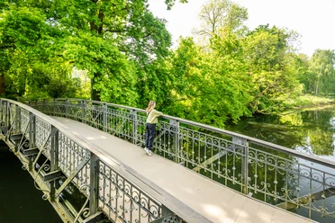 Herford-Aawiesenpark-Teutoburger-Wald-wfg-Herford-D-Ketz-144.jpg Ein grüner Park mit einer verzierten Metallbrücke, umgeben von üppigem Baumwerk und einem ruhigen Teich.