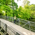 Herford-Aawiesenpark-Teutoburger-Wald-wfg-Herford-D-Ketz-144.jpg Ein grüner Park mit einer verzierten Metallbrücke, umgeben von üppigem Baumwerk und einem ruhigen Teich.