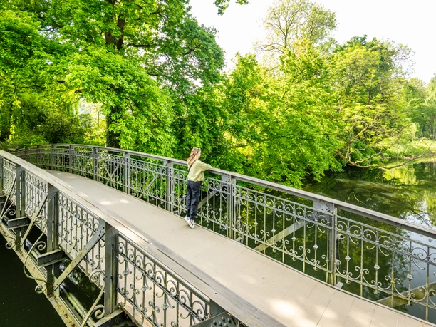 Herford-Aawiesenpark-Teutoburger-Wald-wfg-Herford-D-Ketz-144.jpg Ein grüner Park mit einer verzierten Metallbrücke, umgeben von üppigem Baumwerk und einem ruhigen Teich.