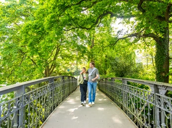 Zwei Menschen spazieren bei sonnigem Wetter über eine Brücke im grünen Aawiesenpark Herford.