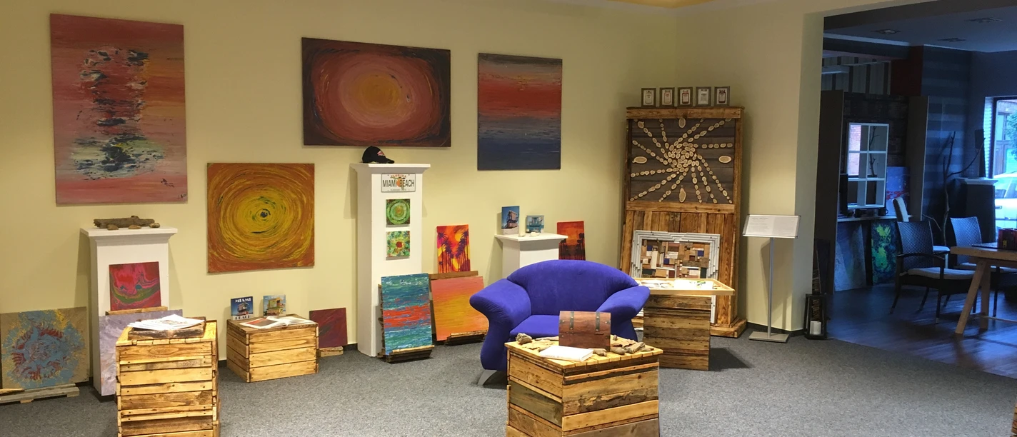 WeserARTelier in Petershagen-Windheim | Ein Ort für Kunst und Kultur Kunstgalerie mit bunten abstrakten Gemälden an cremefarbenen Wänden und blauen Sesseln im Raum.