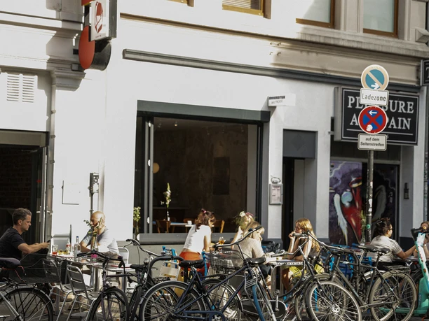 Frohnatur Bistrooo & Bar Bar Bar Straßenszene mit entspannten Menschen beim Cafébesuch und zahlreichen Fahrrädern im Vordergrund.