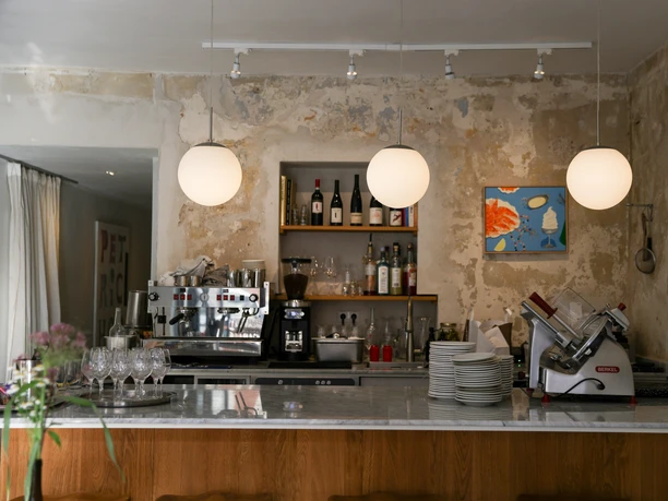 Frohnatur Bistrooo & Bar Bar Bar Drei kugelförmige Hängelampen leuchten über einer Theke mit Weingläsern, Kaffeemaschine und Brotschneidemaschine.