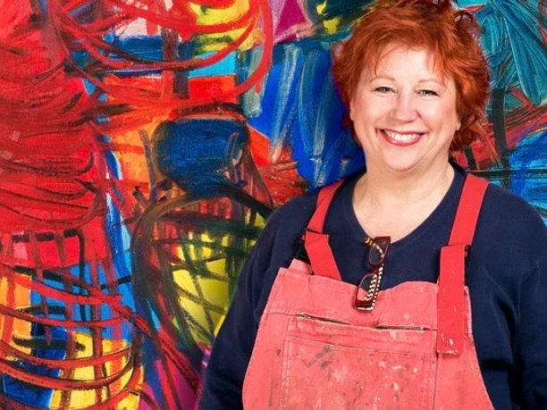 Barbara Salesch steht lächelnd vor einem gemälde mit intensiven Rot- und Blautönen und trägt eine rote Schürze.