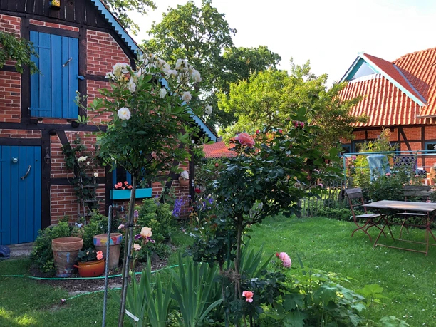 Gepflegter Garten mit roten Ziegelgebäuden, üppigen Blumen und grünen Rasenflächen im Sonnenlicht.