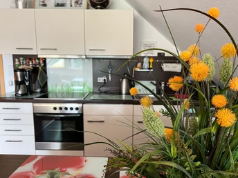 Küchenzeile der Ferienwohnung Naturblick In einer Küche mit gelben Blumenarrangement im Vordergrund, ist der eine Kaffeemaschine, ein Wasserkocher auf der Ablagefläche neben dem Herd inkl. Backofen platziert sind.