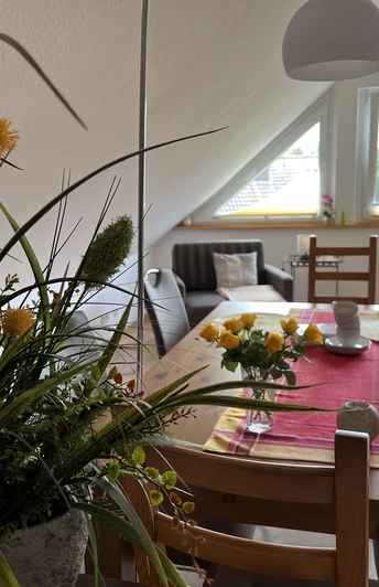 Essecke der Ferienwohnung Naturblick Das Bild zeigt ein Wohnzimmer mit einem großen Esstisch und dekorativen Blumen auf einem Tischläufer.