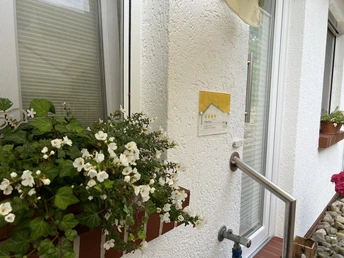 Eingang der Ferienwohnung Naturblick Weiße Petunien schmücken ein Fensterbrett vor einer strukturierten weißen Wand. Ein gelbes Schild neben dem Fenster zeigt vier Sterne. Ein Handlauf aus Metall zieht sich am Fenster entlang.