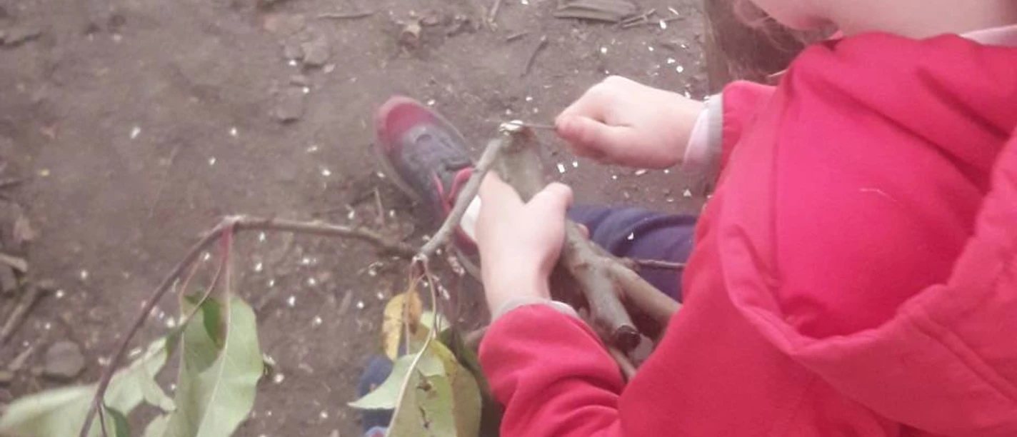 Herbstkurse_254_Kind_schnitzt_C_Lucia Gruber.jpg Kind schnitzt an einem Ast im Freien, umgeben von Laub und Holzstücken am Boden.